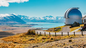 A silver astronomical observatory dome sits on a rugged, tussock-covered hill. In the background, a large bright blue lake stretches toward a majestic range of snow-capped mountains under a sunny sky with scattered clouds.