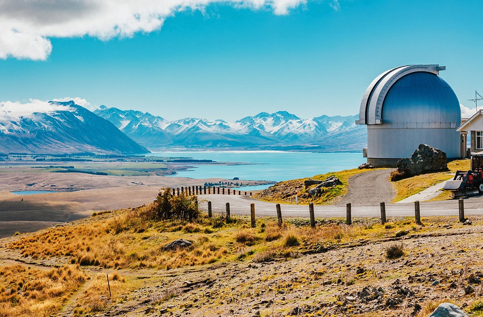 A silver astronomical observatory dome sits on a rugged, tussock-covered hill. In the background, a large bright blue lake stretches toward a majestic range of snow-capped mountains under a sunny sky with scattered clouds.