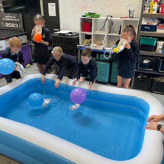 A group of primary school students standing around a blue inflatable pool indoors. They are testing small toy boats powered by balloons in the water, participating in a hands-on science or engineering experiment.