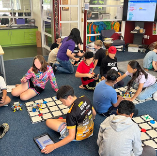 Several students sitting on the floor of a classroom working with small programmable robotics on grid mats. One student uses a tablet to code or control a robot, highlighting an interactive STEM robotics lesson.