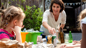 An educator guiding young students through a place-based sustainability and waste management lesson with recycling containers in a school garden.