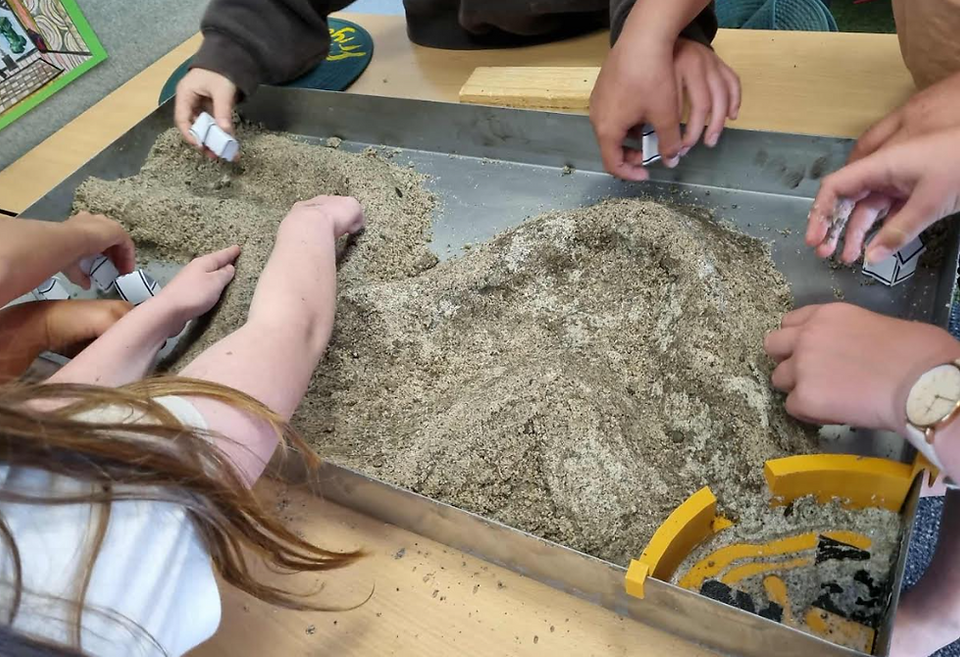 A close-up shot of students' hands interacting with a large metal tray filled with wet sand. They are shaping the sand and using small tools, likely participating in an earth science or geography-related topography experiment.