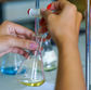Hands using a pipette to transfer liquid into a flask, in a lab setting. The flask contains a yellow solution.