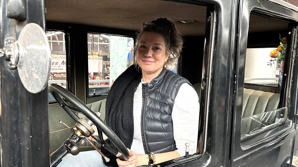 Lächelnde Frau sitzt am Steuer eines alten schwarzen Autos in einer Halle.