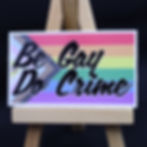 3" wide sticker of progress pride flag with black text: "Be Gay Do Crime" on mini easel