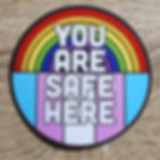 Circle sticker with a rainbow on the top half, trans pride colors on bottom, "You Are Safe Here" in white text