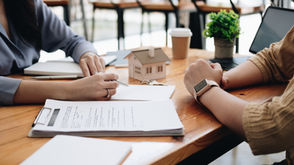 Two people review real estate documents at a table with a house model, keys, and a laptop in view.