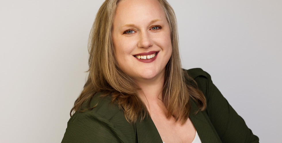 Professional headshot of a confident woman with light blonde hair, wearing a green blazer and a warm smile, captured in a studio setting for personal branding.