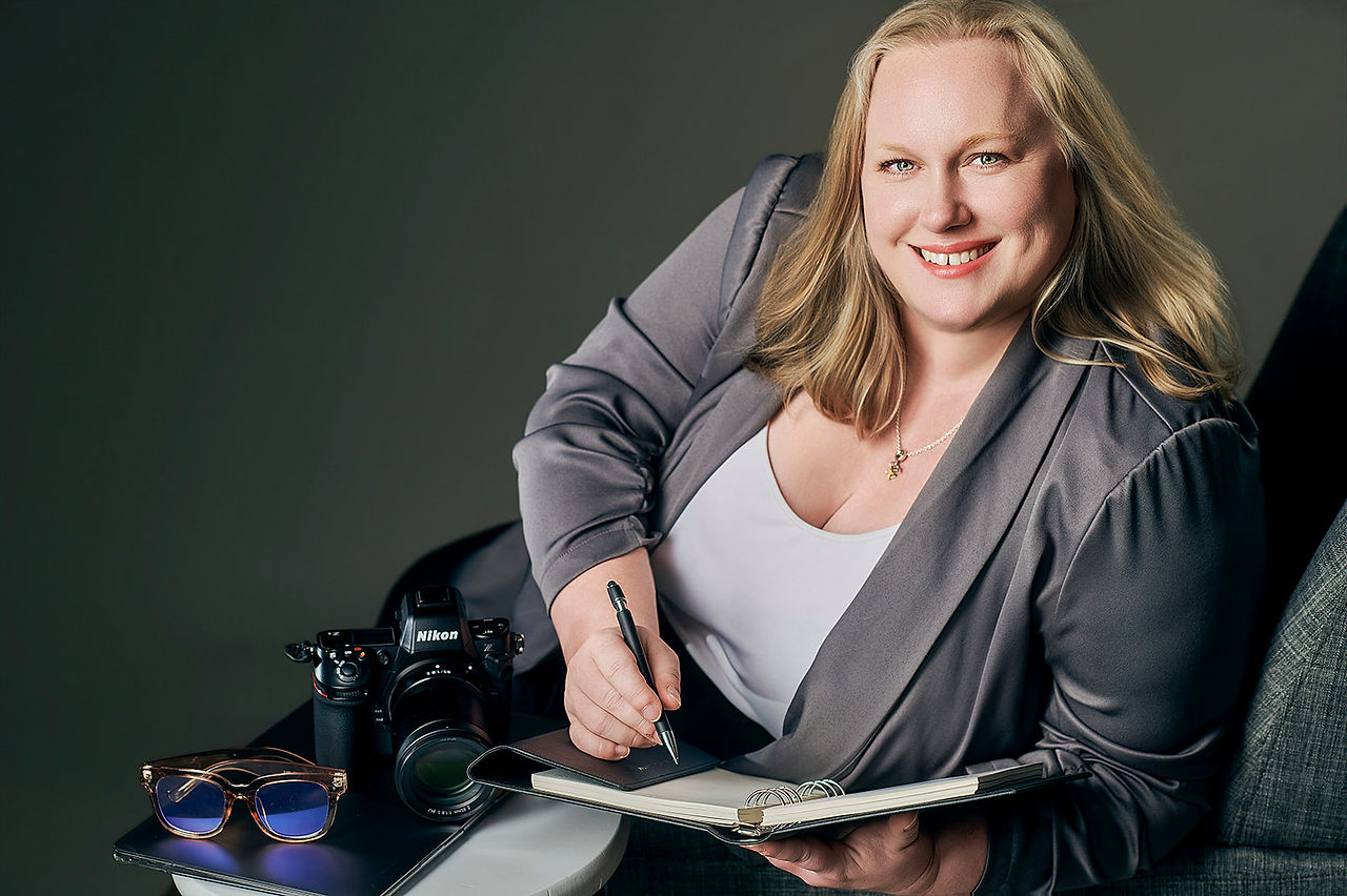 Amanda seated in the studio with a soft smile, posing with a camera and wearing a slate blazer.
