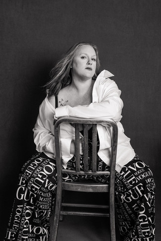 Empowering portrait of a woman seated backward on a chair with wind-swept hair, wearing statement pants and a white shirt, gazing upward in determination.