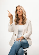 Seated studio portrait of a healthcare provider in a white blazer pointing upward.