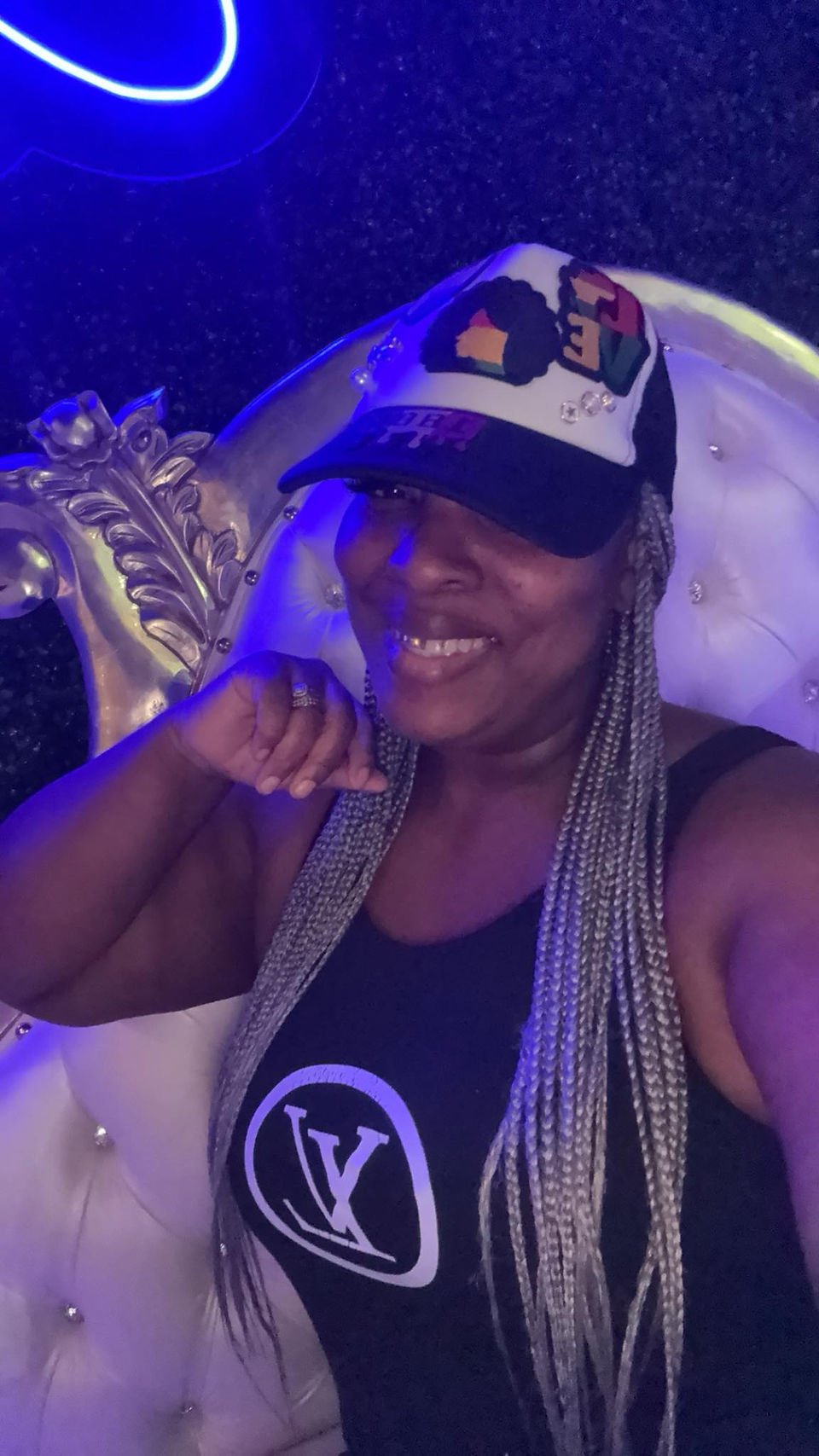 Woman with long silver braids and a colorful cap smiling under neon lights, confidently posing in a lounge chair.