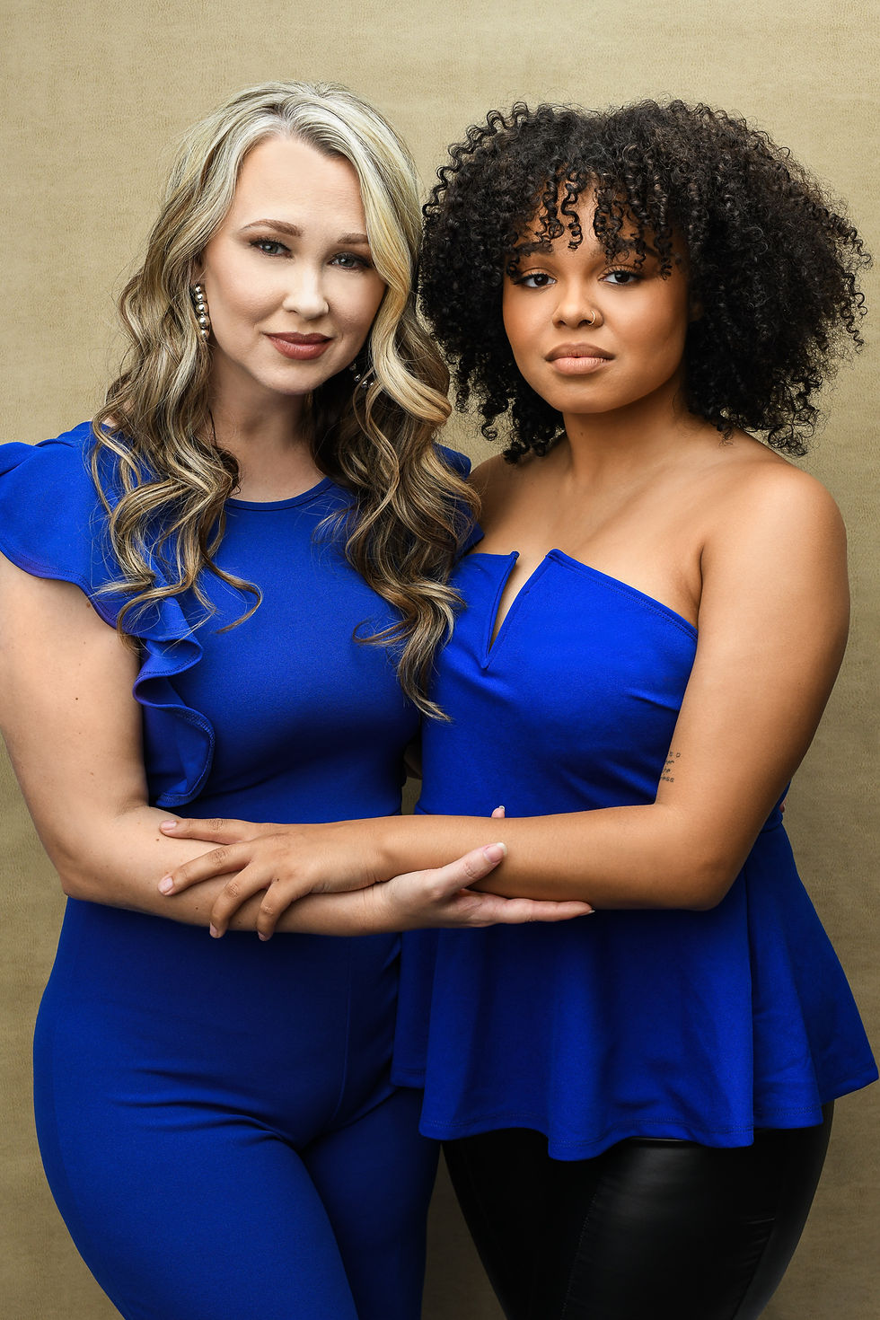 A mother and daughter standing together in elegant royal blue outfits, arms gently linked, with a warm beige backdrop.