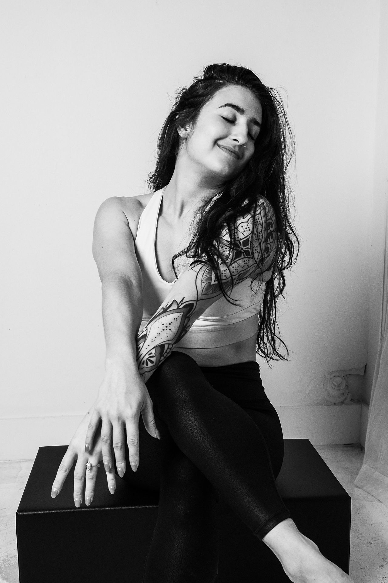 Playful and expressive black and white portrait of a woman sitting on a box, smiling with closed eyes and radiating self-assurance.