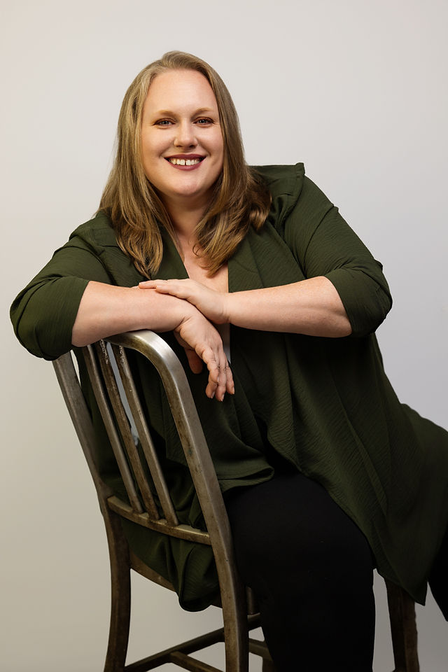 Self-portrait of a woman smiling and sitting on a chair with arms crossed over the back, wearing a green jacket and black pants.