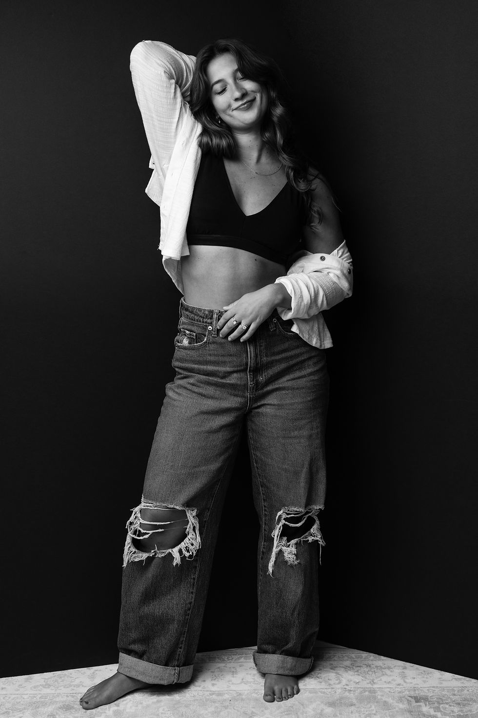 Black-and-white portrait of a young woman smiling softly while posing confidently in a black top, distressed jeans, and an open white shirt against a dark backdrop.