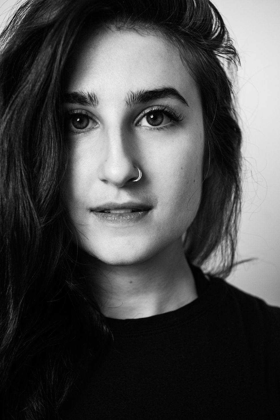 Close-up black-and-white portrait of a woman with natural features, long dark hair, and a nose ring, gazing directly into the camera with a calm and introspective expression.