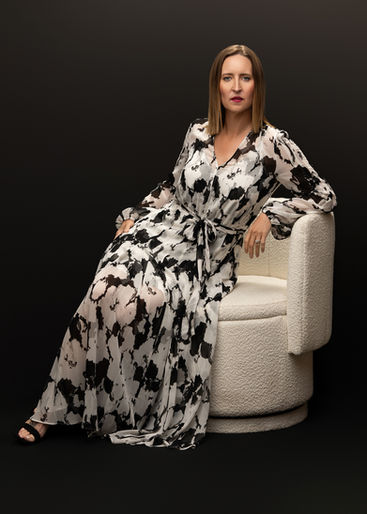 Woman sits elegantly on a white boucle swivel chair wearing a dramatic black-and-white patterned dress, looking poised and powerful against a black backdrop.