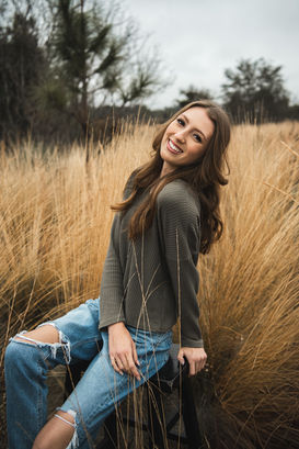 Teen girl sitting in tall grass wearing jeans after styled senior portrait session with Avy Productions