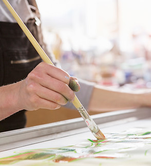 A persons hands holding a paint brush to a piece of canvas as they are painting, the image cuts off so we do not see the entire person nor their painting, just their arms, torso, and hand holding a paint brush