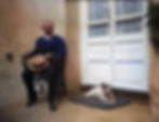 A man sits on a chair and smokes and his dog rests on the bench in front of the door.