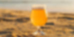 A glass of beer with foam sits on sandy beach at sunset, with golden hues creating a warm, relaxed ambiance.