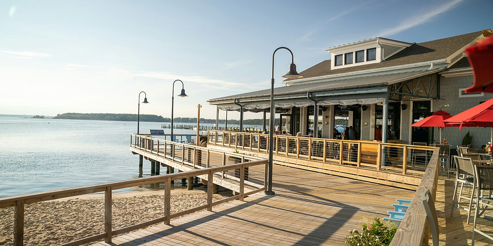 Beachfront restaurant with people dining. Wooden deck, red umbrellas, and ocean view under a clear sky. Relaxed, sunny atmosphere.