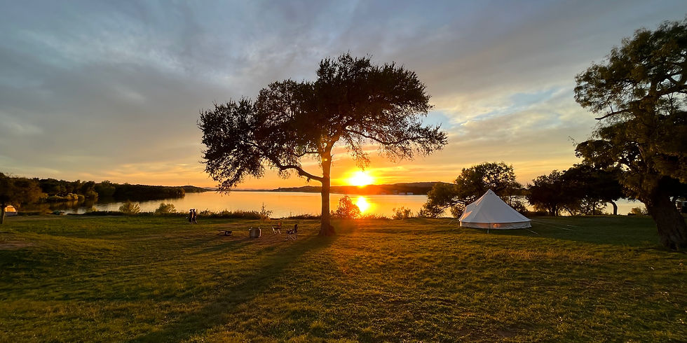 Sunset over a lake with a tree and a white tent on grassy shore. Warm hues dominate the sky; a tranquil, serene atmosphere prevails.