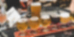 A flight of six beers in small glasses on a wooden tray. Other beer glasses and a menu are on a metal patio table in the background.