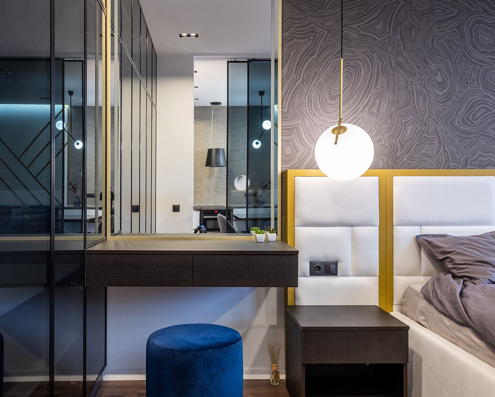 A rectangular mirror in a vertical orientation on top of a bedside table. The mirror has a thin frame made of shiny gold metal with angular geometric shapes trim. The clean-lined minimalist frame contrasts with the soft reflection of the surroundings visible in the mirror's surface. The mirror sits atop a brown vanity table next to a hanging pendant lamp and decor items, adding a touch of glamour to the bedside decor.