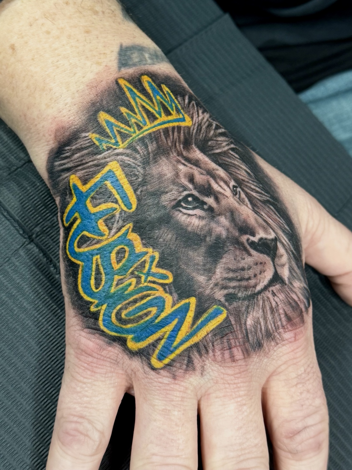 Realistic lion tattoo with blue and yellow King lettering and crown on hand