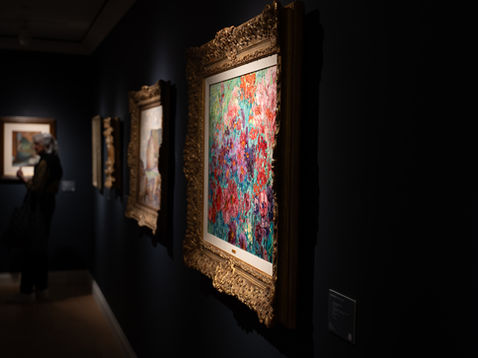 Dark art gallery with ornate gold-framed colorful abstract paintings; a blurred visitor stands viewing them in quiet, moody light