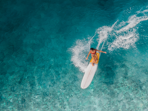 Surfer in red bikini rides a wave on a white board in clear blue ocean. Aerial view with splash trails, evoking a sense of adventure.