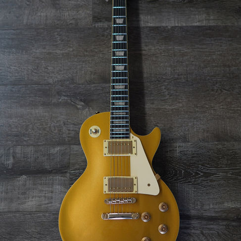 Diagonal high-angle view of a goldtop electric guitar with a cream pickguard and gold hardware on a dark grey wood floor.