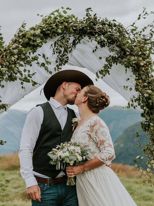 Brudepar som kysser under frodig bue i vakker natur