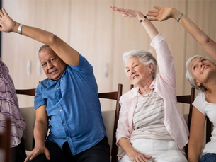 Four older adults sit in chairs and stretch overhead in a bright room, smiling during a gentle exercise class.