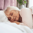 Elderly woman sleeping peacefully in bed, head resting on a white pillow. Soft lighting, pink headboard, and a plant in the background.