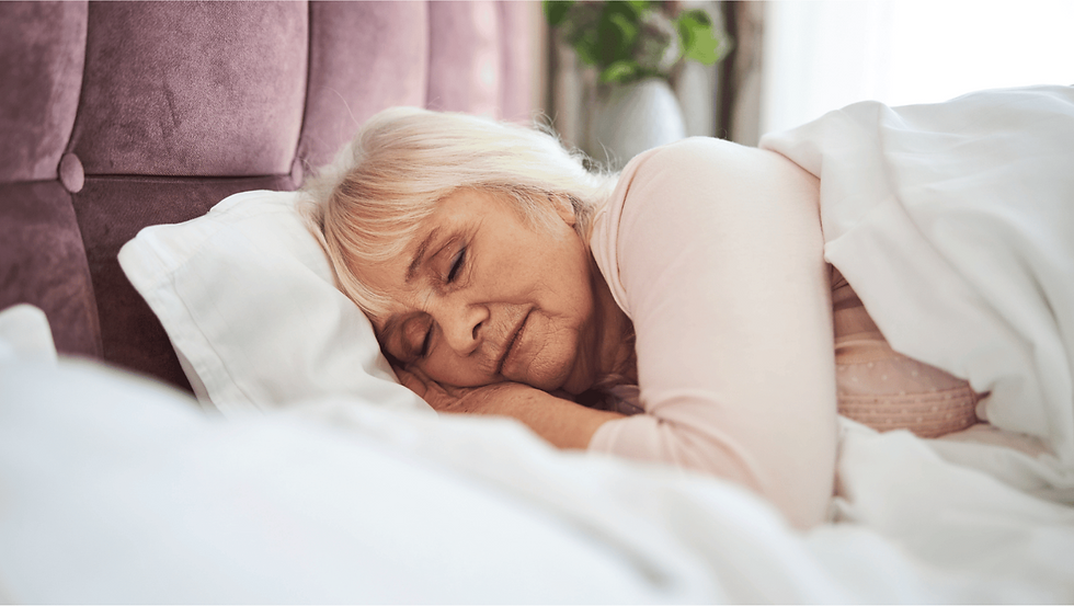 Elderly woman sleeping peacefully in bed, head resting on a white pillow. Soft lighting, pink headboard, and a plant in the background.