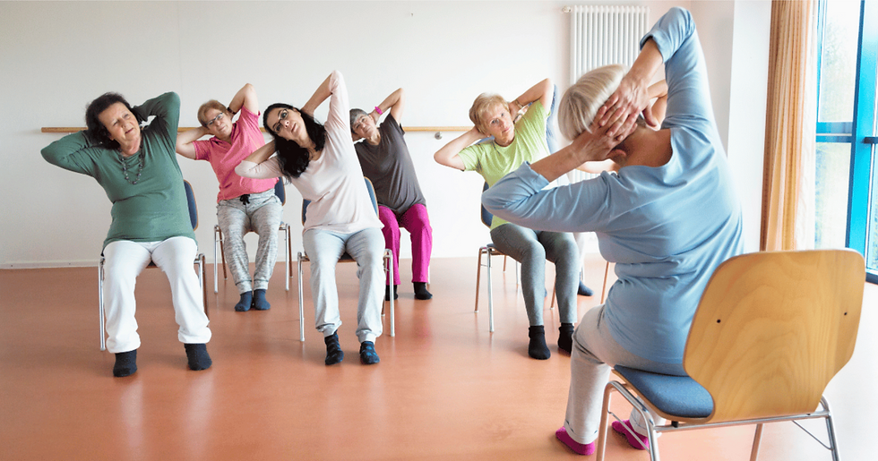 Older adults seated in a yellow room, performing chair exercises. Focused expressions, bright natural light from windows. Comfortable attire.