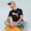 A young man wearing a black Puma T-shirt with a white leaping cat logo, a grey cap, and grey sweatpants. He is sitting on a yellow ottoman with one leg bent, looking to the side with a relaxed pose. His tattoos are visible on his arms