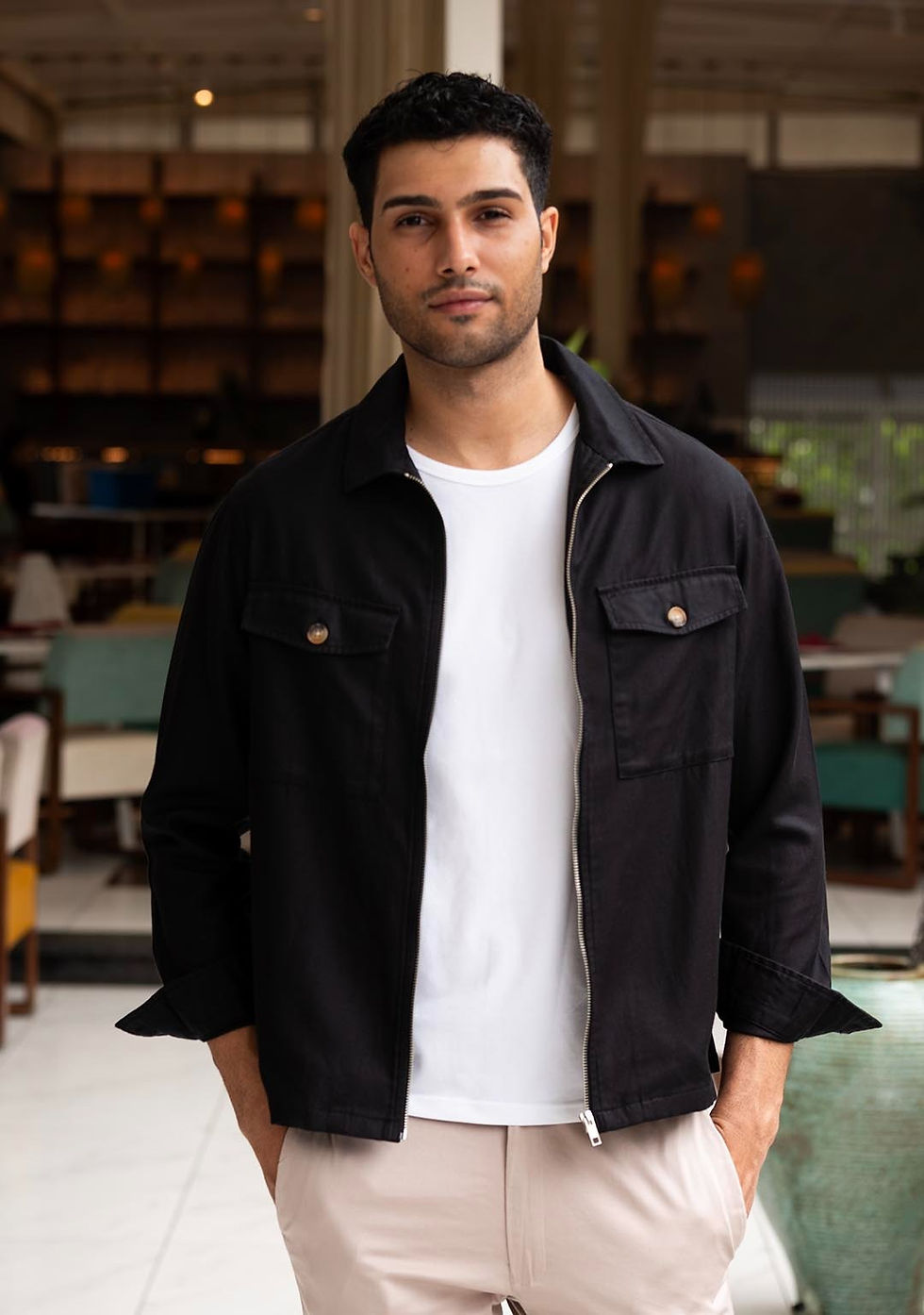 A stylish man wearing a black shacket over a white t-shirt and beige trousers, posing in a modern indoor café setting.