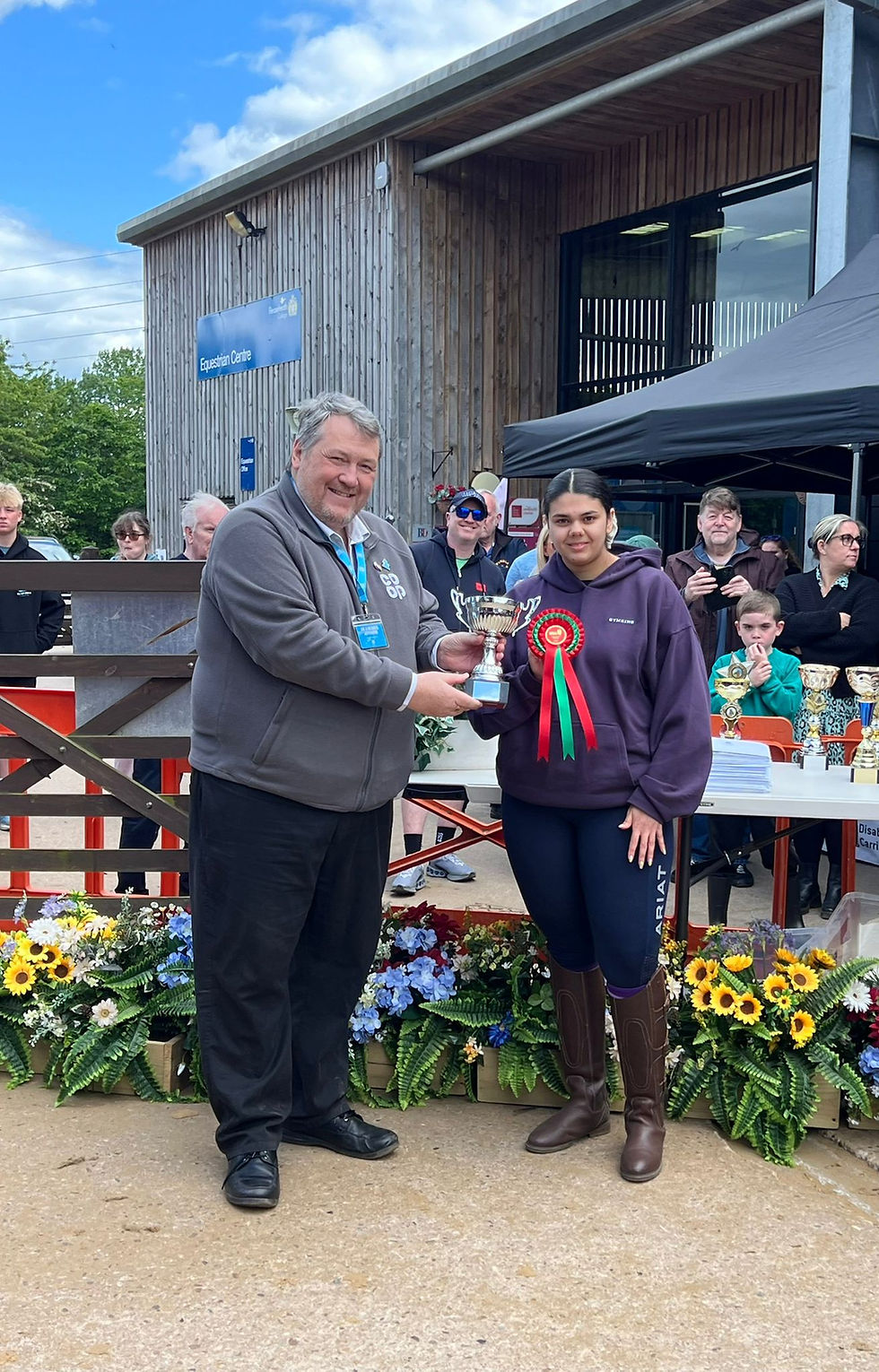 Joyful photo with rider in purple group hoodie being presented with trophy and red rosette.