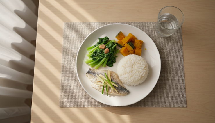 Overhead view of a small balanced meal with fish, rice, soft vegetables, and a glass of water placed aside