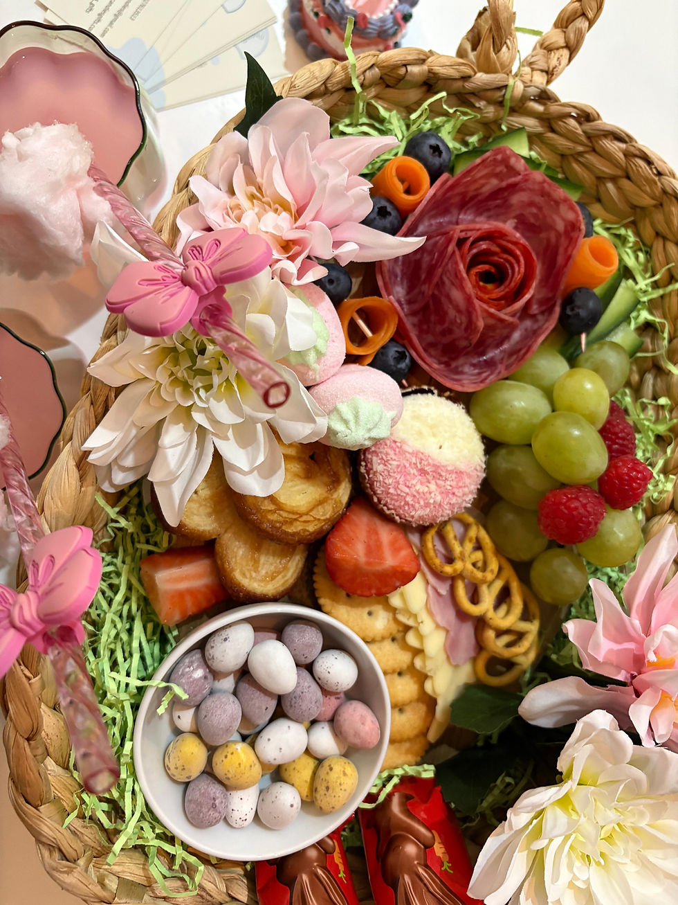Basket with assorted snacks: salami, grapes, berries, eggs, cheese, crackers, flowers. Pink drink with butterfly straw. Bright, festive mood by Creative Candies & Gifts.