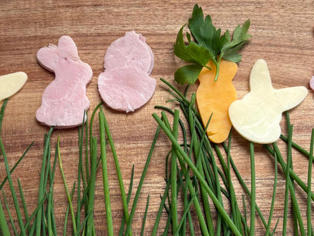 Carrot and rabbit shapes made of cheese, ham, and herbs on a wooden board with green chives below, creating a playful, Easter vibe by Creative Candies.