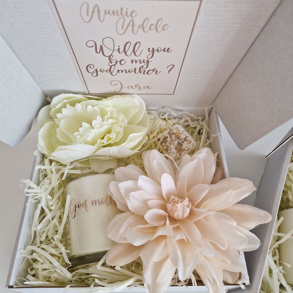 Box with a "Will you be my Godmother?" card, white candle labeled "Godmother," and two large pale flowers on shredded paper by Creative Candies & Gifts.