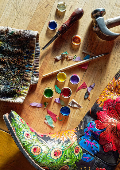 Hand-painted custom leather cowboy boot with peacock feather and floral motifs on a craftsman's workbench.