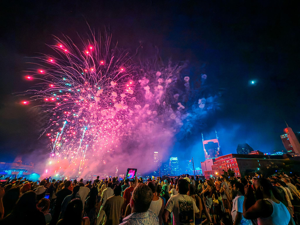 A crowd watches vibrant red and pink fireworks over a city skyline at night. The scene is festive, with people capturing the moment.