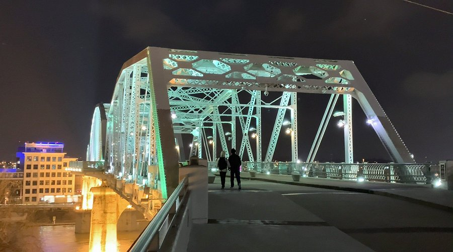Two people walk on an illuminated steel bridge at night, with city lights visible in the background, creating a serene atmosphere.