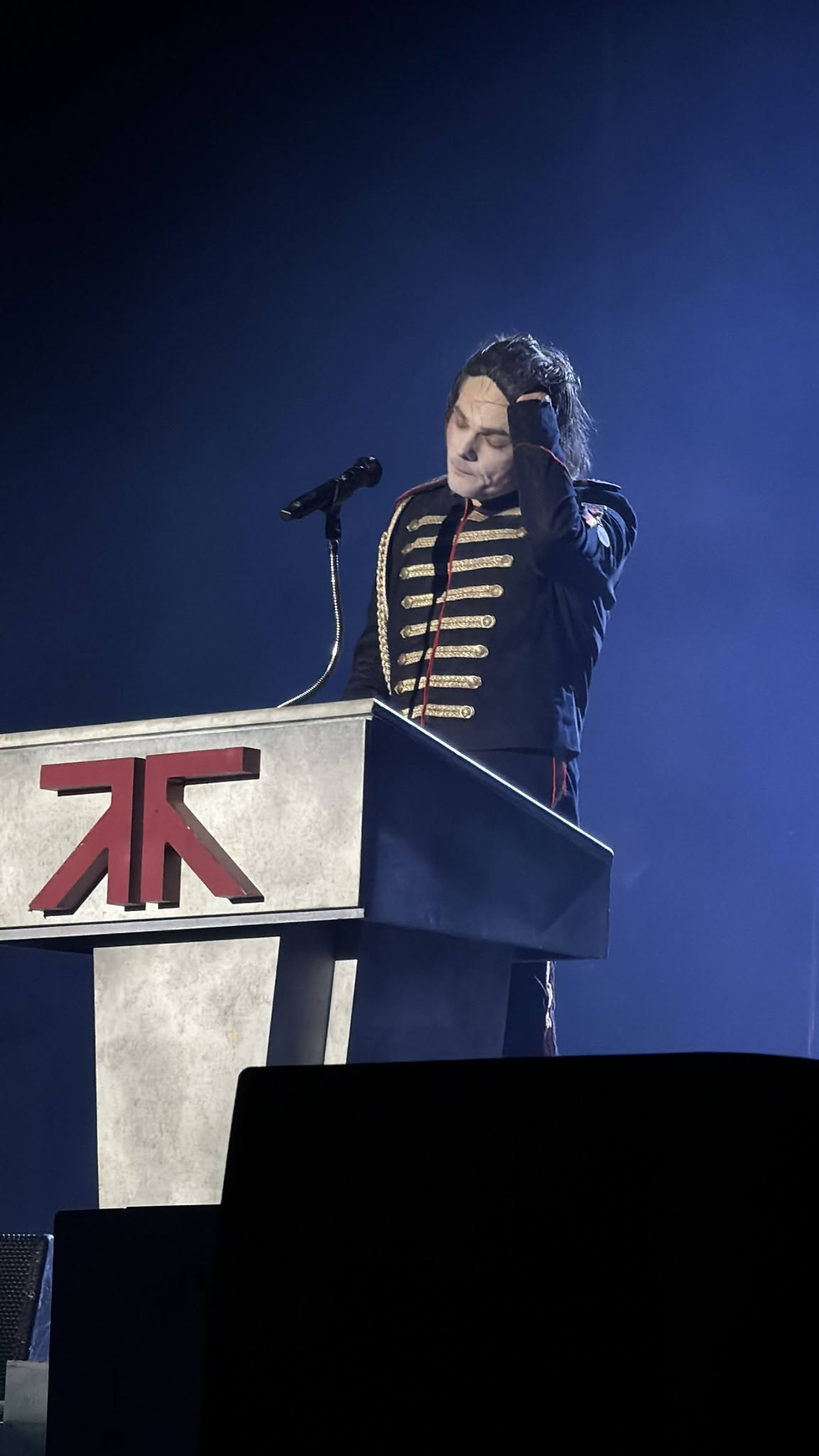 Michael Jackson impersonator in a military jacket stands at a podium with a microphone, hand on head, under blue stage lights; red TK logo